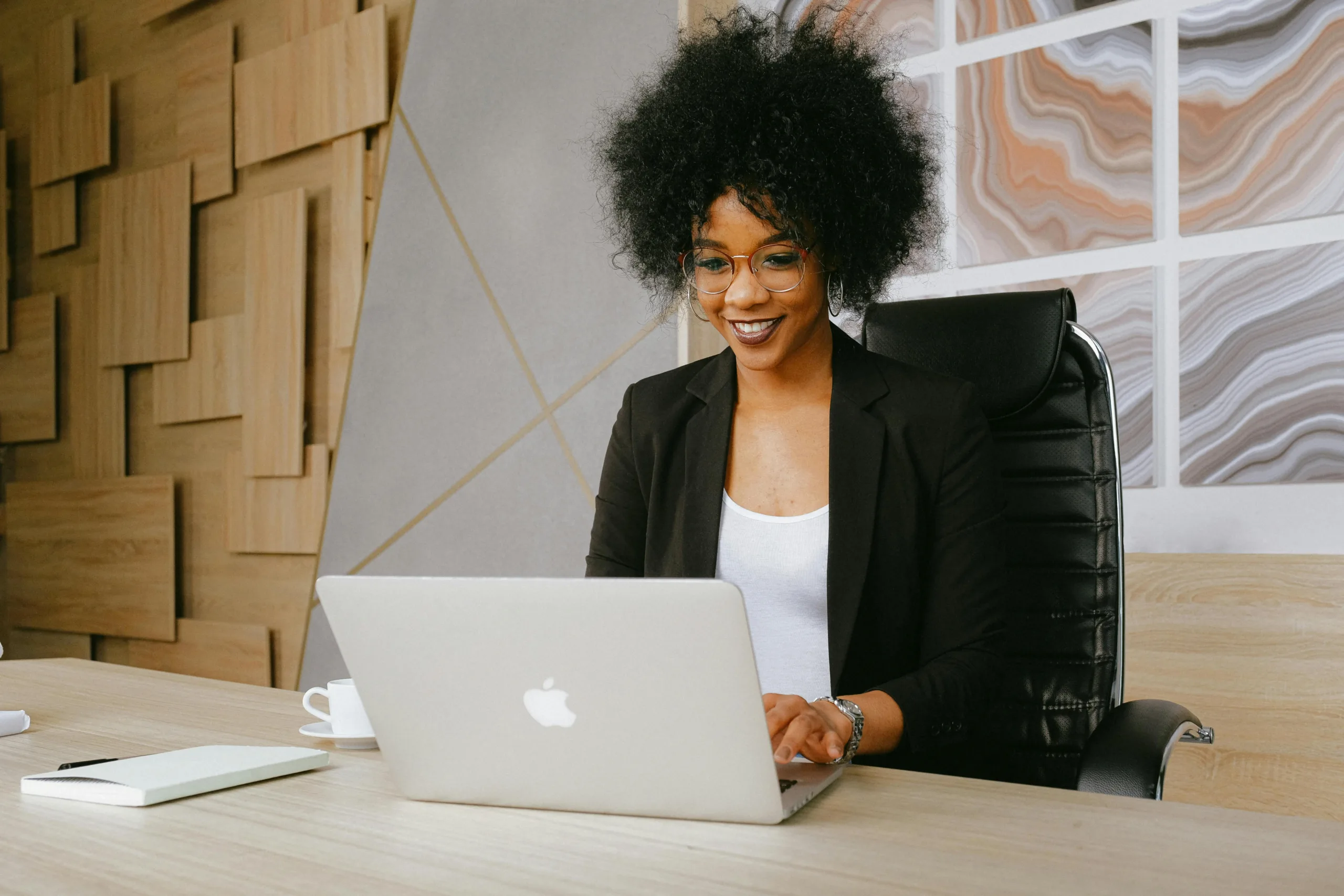 Black woman in professional apparel smiling and sitting at a desk in front of laptop.