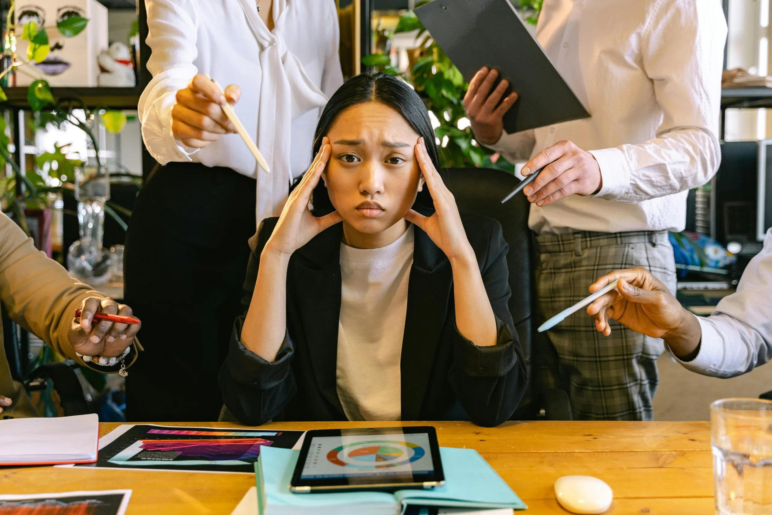 Overworked female employee holding her head with both hands looking at viewer in discomfort, with people surrounding her out of view pointing at her displeased.