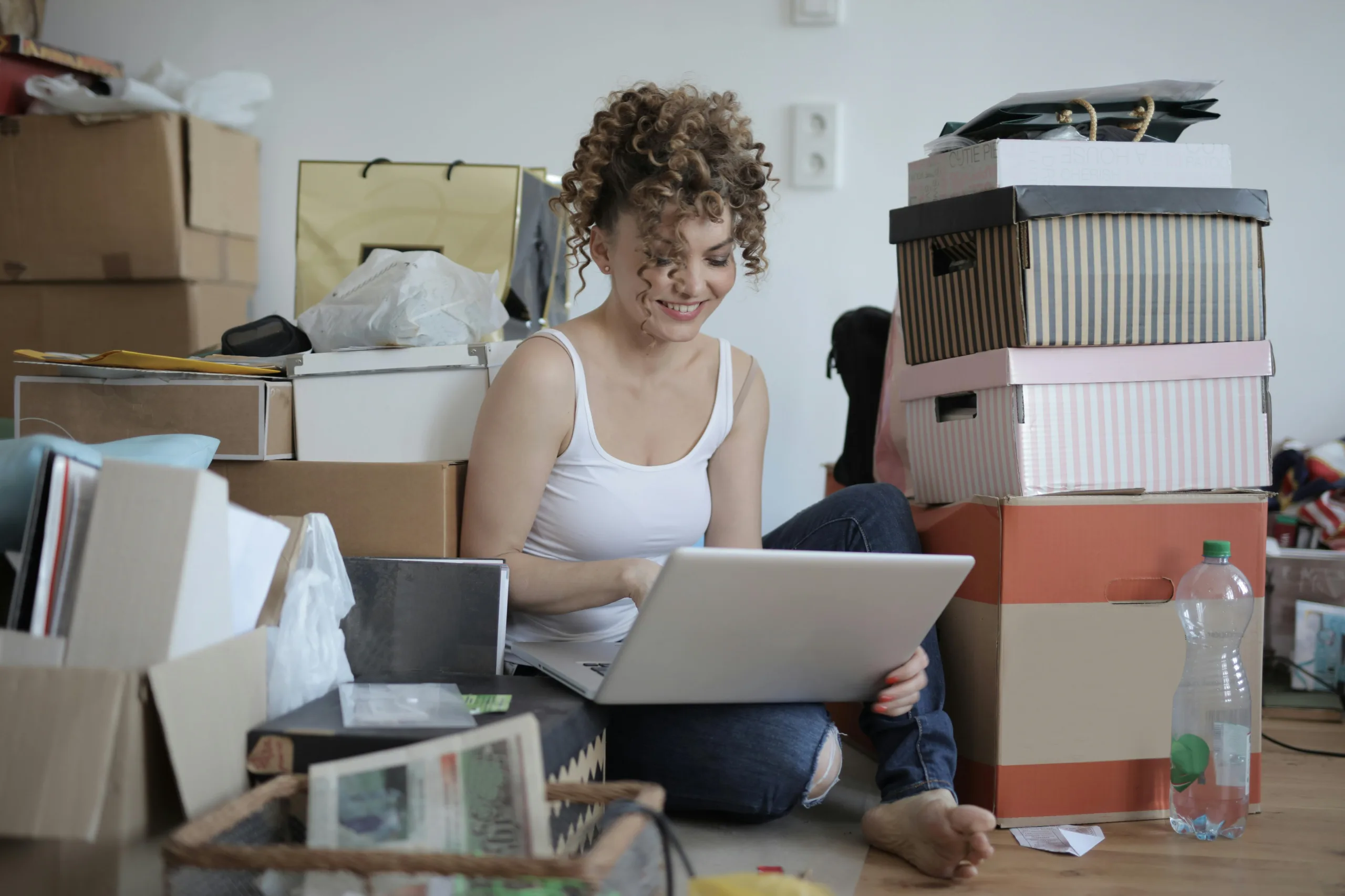 Curly haired woman sitting beside her clutter, smiling at her laptop ignoring the mess around her.